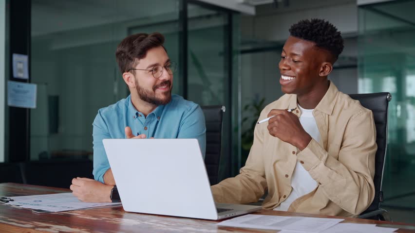 Two busy diverse male employees using laptop computer talking in office. Professional young business men workers team having conversation working on project sharing ideas sitting at work desk. - Powered by Shutterstock - Get 15% off with code: PIKWIZARD15