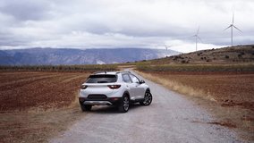 Rear view of a car on a dirt highway moving away, driven on a landscape with windmills on the background. Wide shot with copy space.