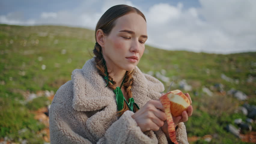 Woman peeling apple fruit on outdoor journey. Closeup mountain traveler having juicy healthy snack looking camera. Thoughtful girl with braids rest on green hill alone. Eco friendly sustainable living