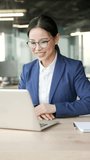 Vertical video. Asian businesswoman talking on a video call using laptop sitting at workplace in office. Woman in formal suit communicates at online conference or chats remotely with business partner