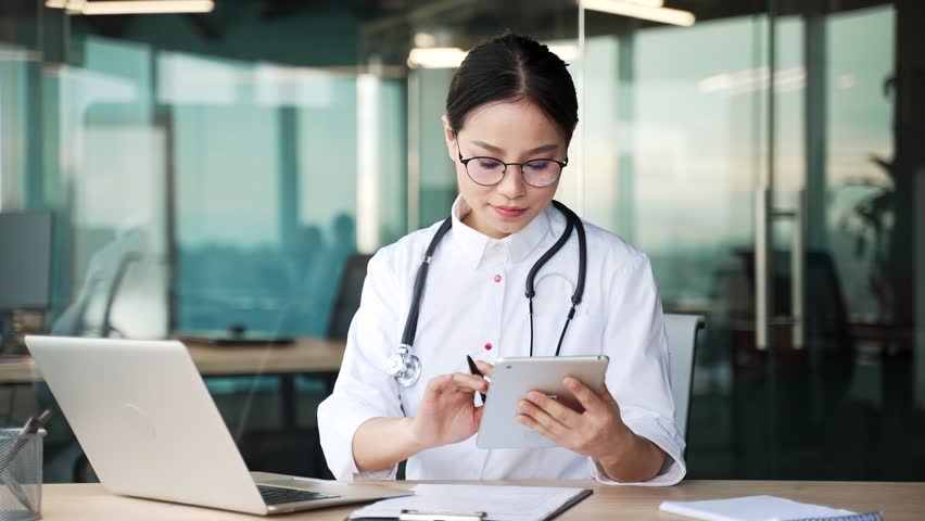 Young asian female doctor works using a digital tablet and laptop while sitting at workplace in a modern hospital clinic. Woman medical worker physician in a white coat is busy with a report in office