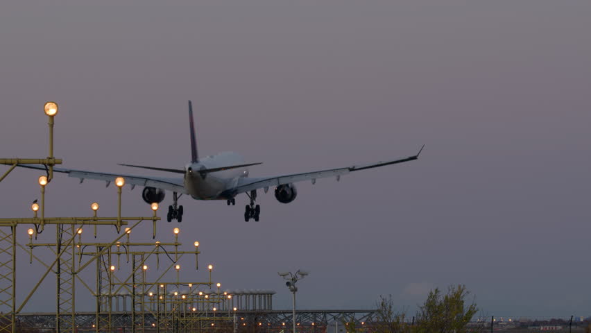 Big commercial airplane with extended landing gear approaching runway during evening, with runway lights visible