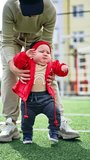 Unrecognized woman helping the toddler to walk. Kid in red jacket learning to walk by the soft covering of stadium. Vertical video.
