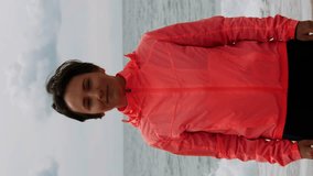 Portrait of smiling woman in orange jacket standing at edge of beach. Vertical video. Sportive female standing on beach at stormy weather