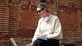 Successful female student having online classes through modern laptop on fresh air. Talkative caucasian woman with sunglasses and wireless headphones on neck making notes in copybook outdoors.