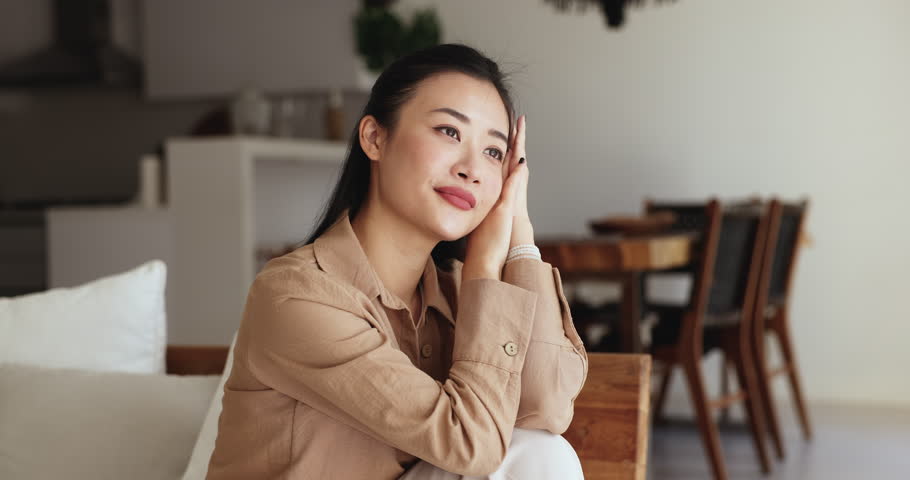 Close up shot dreamy pretty Asian female enjoy carefree relaxation seated on comfortable sofa in contemporary living space, laugh, look at camera, posing, feel happy alone at own or rented apartment Royalty-Free Stock Footage #3484629793