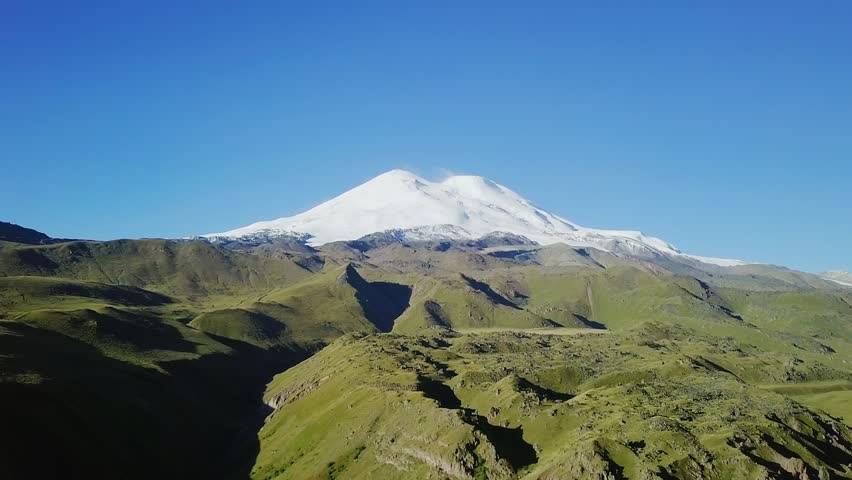 Elbrus from the north in the summer. A dormant stratovolcano with twin snow-capped summits rises above rolling green alpine meadows and deep glacial valleys under a clear blue sky. The northern approach reveals an untouched high-mountain landscape of grassy ridges, volcanic rock outcrops, and steep ravines carved by meltwater streams, with the massive ice-covered peak dominating the horizon. The scene captures the raw scale of Europes highest mountain as viewed from the less-traveled northern route, where subalpine terrain transitions toward the glaciers and snowfields of the upper slopes. Bright midday sunlight casts deep shadows across the undulating terrain, emphasizing the rugged texture of the Caucasus foothills. The wide aerial perspective showcases the dramatic elevation gradient from verdant valleys to the permanent snowline, a classic summer vista in the Greater Caucasus range, Point of interest