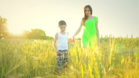 Mother and her son walk across field with green wheat in rays of hot sun