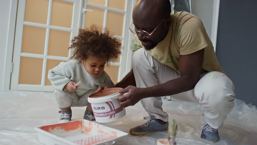 Handheld medium footage of Biracial little girl watching her African American dad pour beige paint from bucket into tray before painting walls in new apartment Royalty-Free Stock Footage #3484967837