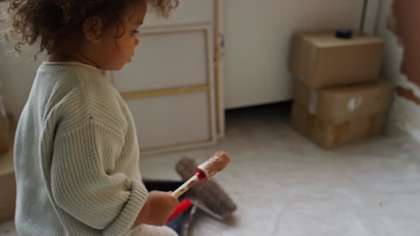 Tracking side footage of little curious Biracial baby girl with curly hair coming up to wall with paintbrush and painting it with brown color during renovation process Royalty-Free Stock Footage #3485030569