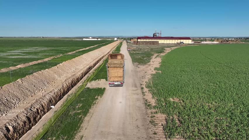 Aerial video following a truck loaded with straw bales along a dirt road between agricultural fields with a large ditch next to the road. Agricultural trade concept