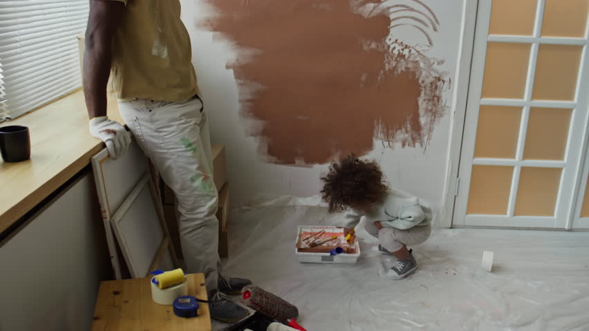 Side tilt footage of single African-American dad watching his little Biracial daughter painting walls brown using paint roller while renovating room into nursery in new apartment Royalty-Free Stock Footage #3485043787