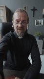 Vertical handheld POV shot of mature Catholic priest wearing earbuds sitting in office holding smartphone talking on video call