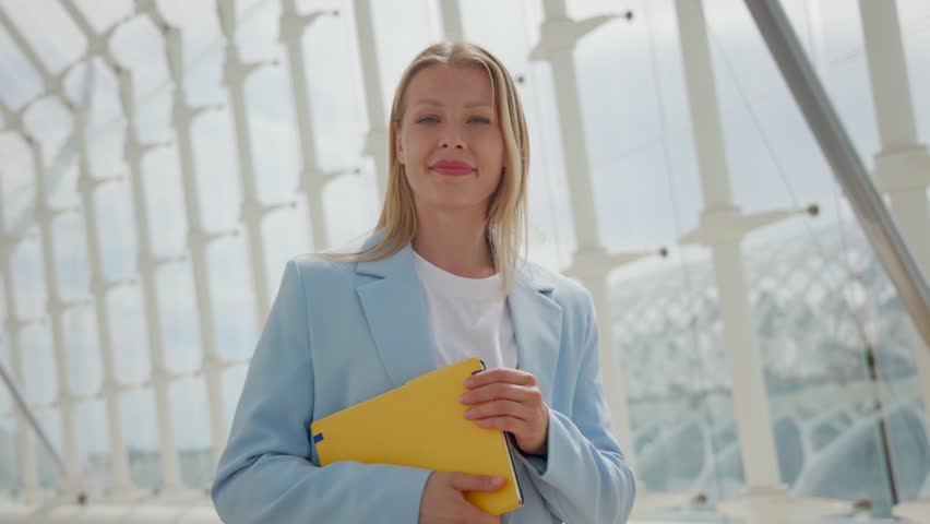 Serene blonde businesswoman in a professional light blue suit confidently holding a yellow document folder, standing with white geometric columns in the soft-focused background. Slow motion.  Royalty-Free Stock Footage #3485094745