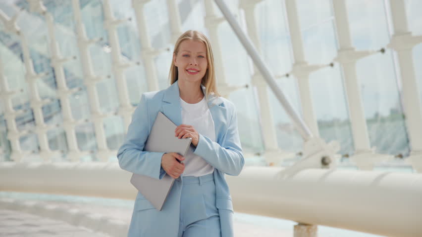 A poised and smiling blonde confident businesswoman carrying a laptop, dressed in a smart light blue suit, walks confidently with architectural structures in the blurred background. Slow motion.  Royalty-Free Stock Footage #3485095141
