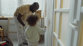 Side handheld footage of cute little daughter with curly hair helping father paint wall with little brush drawing abstact doodles while dad using paint roller - Powered by Shutterstock - Get 15% off with code: PIKWIZARD15