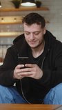 Vertical video. Young man sitting on sofa reacts to what sees, surfing internet on mobile phone, checking email, reading media news, scrolling social medias, using mobile applications on smartphone at