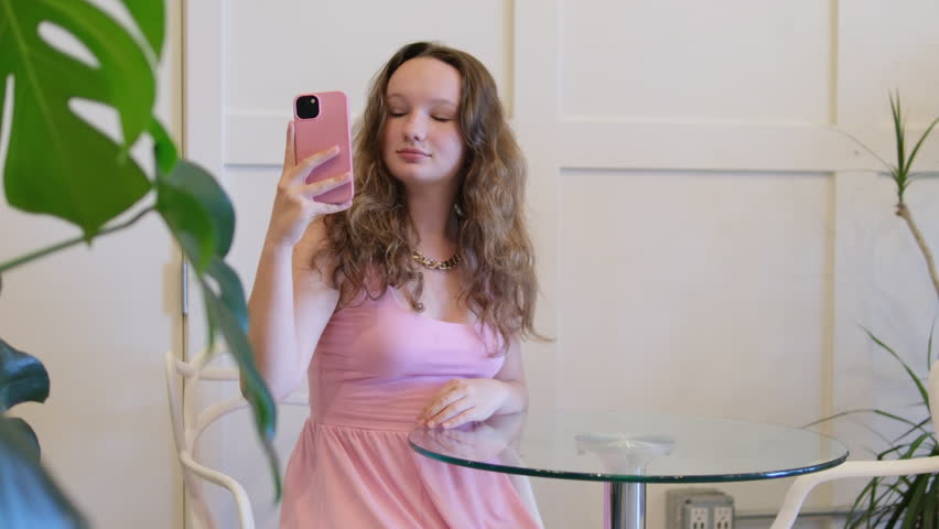 Straighten your hair. girl in a pink dress takes a selfie in a cafe at a glass table against the background of a white wall and palm leaves