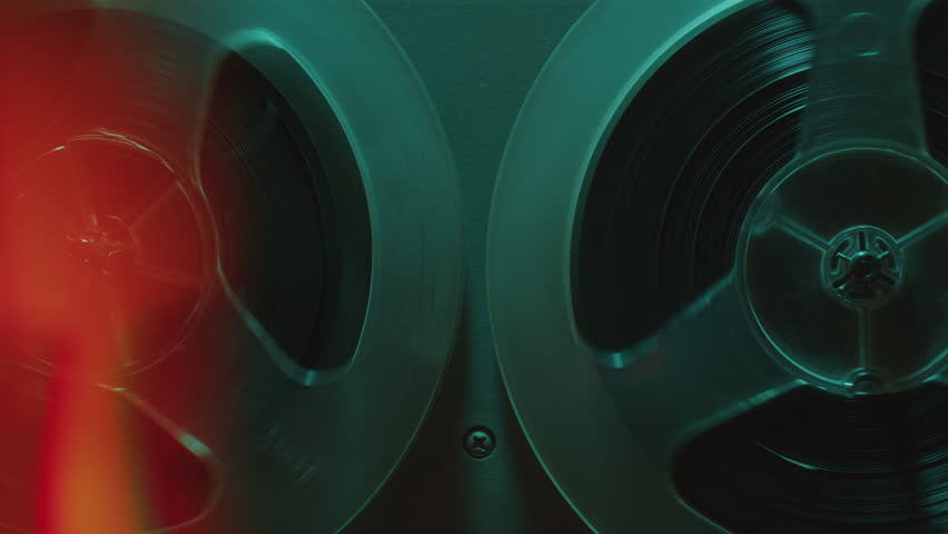 Retro-styled open reel tape recorder with rotating spools in dark studio with blue neon light. Zoom off, close-up shot