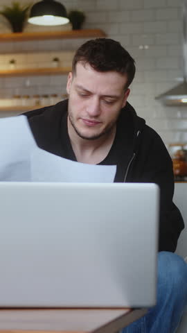 Vertical video. Young man pay bills with laptop, use calculator count expenses, make budget control, manage finances, review overdue loan payments feel stressed while sitting on sofa at home.