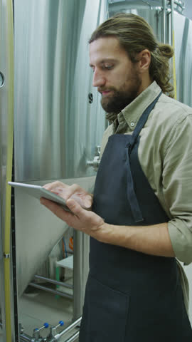 Medium vertical shot of young Caucasian man in apron, with red hair, beard and moustache standing by beer fermenter tank, tapping on pressure gauge and filling out checklist on tablet computer