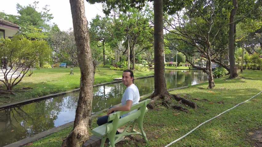 The young man relaxing by the river. He is sitting on the green bench and lookin around. He enjoys the day at the amazing park.