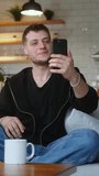 Vertical video. Smiling man sitting on sofa holding mobile phone, waving and greeting while having video conference call on smartphone. Man looking at screen, talking, smiling, enjoying online chat at