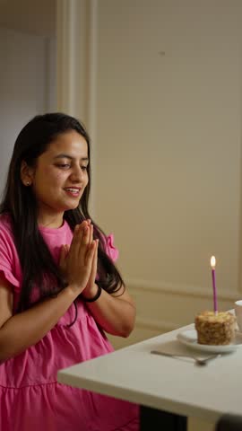 A happy brunette girl in a pink dress sits in front of a table in front of a small cake and blows out a pink candle on it applauding and clapping her hands in a modern apartment