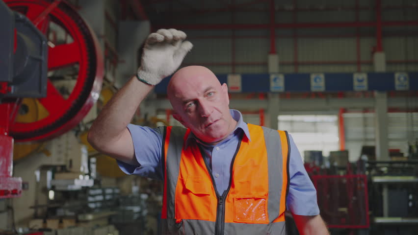 A hardworking industrial worker takes a brief pause to wipe sweat from his brow, reflecting the effort and dedication in a busy factory setting, Slow Motion