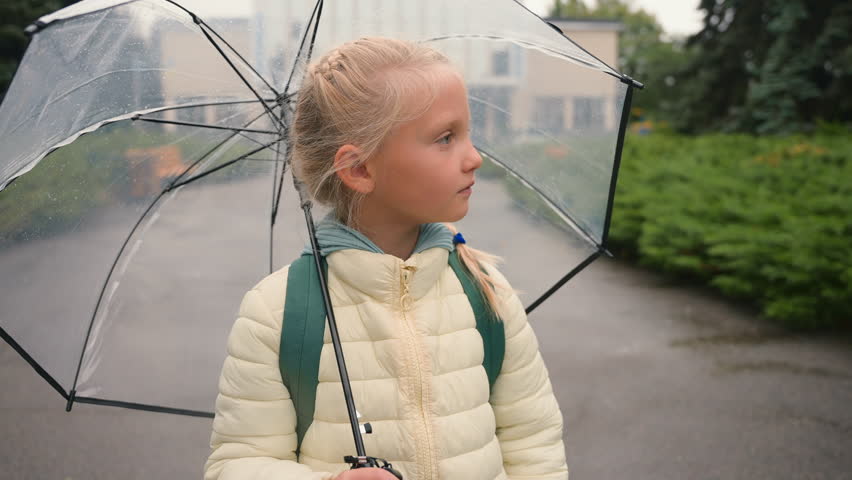Little European girl looking at camera smiling holding umbrella rainy weather child pupil kid city outside park expression funny childish parasol backpack primary school kindergarten preschool enjoy Royalty-Free Stock Footage #3485568029