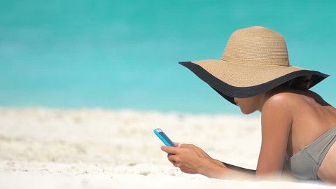Woman using phone app on beach on summer travel vacation sunbathing on sand. Young female on smartphone lying on beach towel wearing sunhat by sea. She is wearing bikini while relaxing during summer.