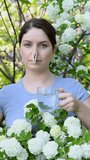 Caucasian woman takes an antihistamine medicine and removes a clothespin from her nose near a flowering tree. Vertical video. 