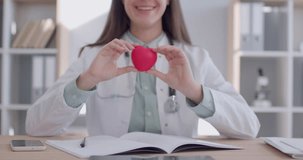 Close up portrait of a female smiling woman doctor cardiologist sitting at the desk on her workplace holding red heart in medical clinic. Health care, cardiology concept. 4K video. Slow motion video.