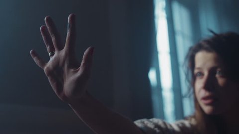 Helpless blue-eyed woman reached out her hand as trying to grab something. Dark room, moody atmosphere, paranormal activity. Morning sun shining through the window