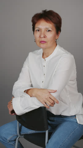 Vertical medium portrait of mature short haired woman smiling at camera while sitting on chair in studio on clear grey background