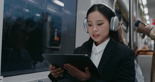 Beautiful Asian woman sitting next to window while watching video or movie on large tablet device. Listening to music in headphones. Tapping on screen and beginning to smile. Public transportation.