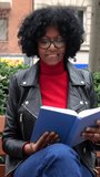 Vertical video. African American middle aged woman reading a book sitting on a bench in a city