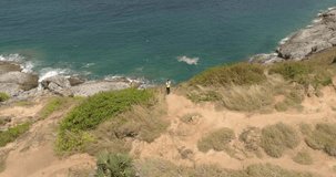 Video from drone. Sportive beautiful girl standing between rocks at cliff washed by sea on a sunny day. The woman stay between rocks. Hiking. Outdoor taining. Nai Harn cliff, Phuket island, Thailan.
