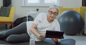Old woman using digital tablet pc and drinking water from bottle resting on mat at home