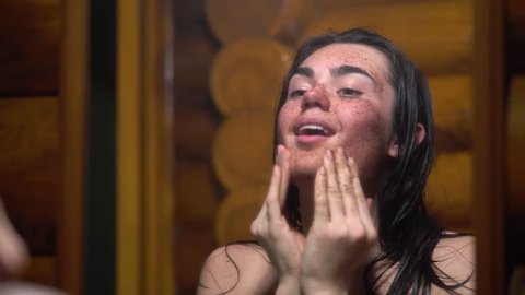 Brunette young girl after sauna happy and smiling applies coffee scrub on her face looking in the mirror