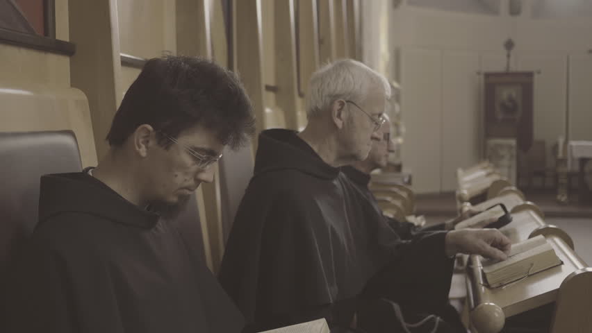 Monks reading Bible together while sitting in church side view medium shot. Christian monks praying liturgy with psalmbook. Liturgy of the Hours concept