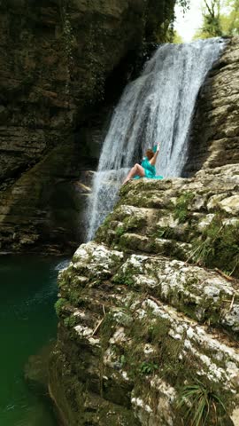 Beautiful girl in a long bright fabric poses against the background of a magnificent waterfall among the mountains. 4K vertical video