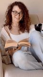 Smiling curly hair woman with glasses reading book sitting on sofa speaking with her dog. Weekend relaxed hobby day. Vertical video footage 