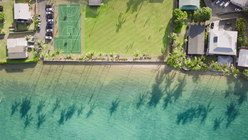 Overhead view of Oahu revealing a shoreline soccer field bordered by palm trees, with their shadows dancing on the turquoise sea. Footage 4K.  Royalty-Free Stock Footage #3486415747