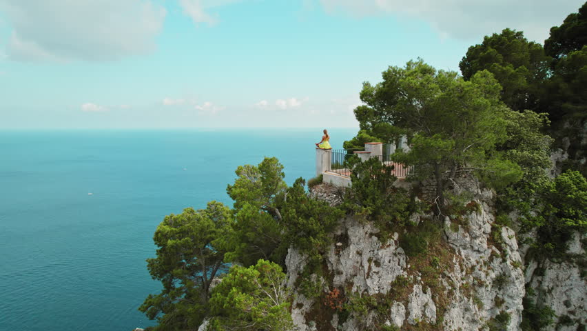 A woman admires the view of the Faraglioni stacks in Capri, Italy from a cliffside. Observation from the summit on breathtaking landscape. Royalty-Free Stock Footage #3486511375