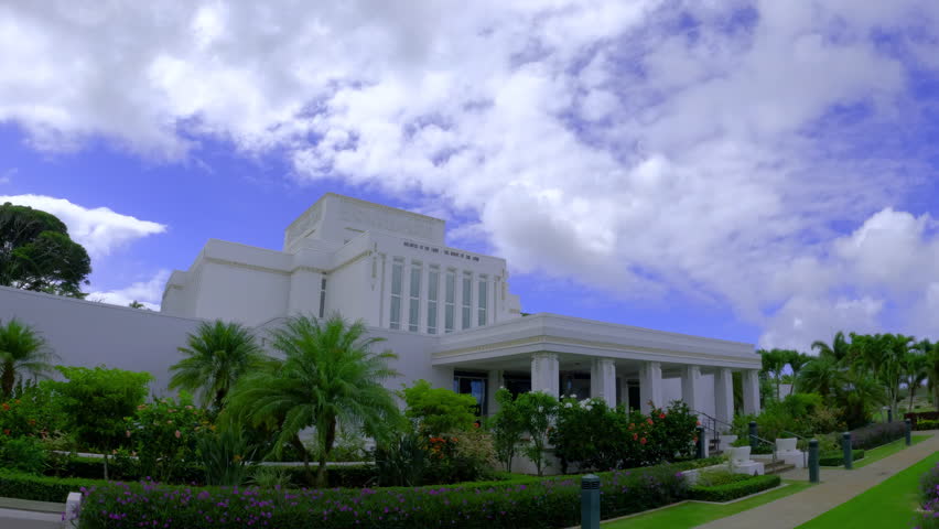 Side view of Laie Hawaii Temple surrounded by flowers and greenery. Establishing shot of white Mormon church on a cloudy day.
