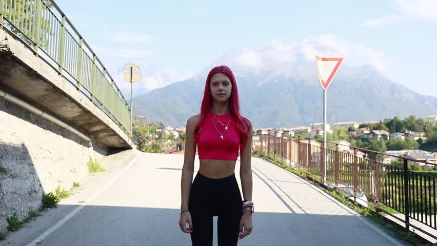a young woman smiles at the camera that accompanies her as she comes toward us, moving forward (relative to herself) walking down a street in a small town in Europe that lies between the mountains