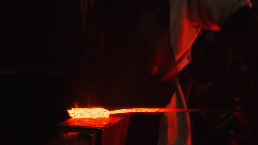 A blacksmith uses a hammer to shape a piece of red-hot metal in a forge on an anvil, creating a future product, sparks fly from blows. The process takes place in the darkness of the forge.