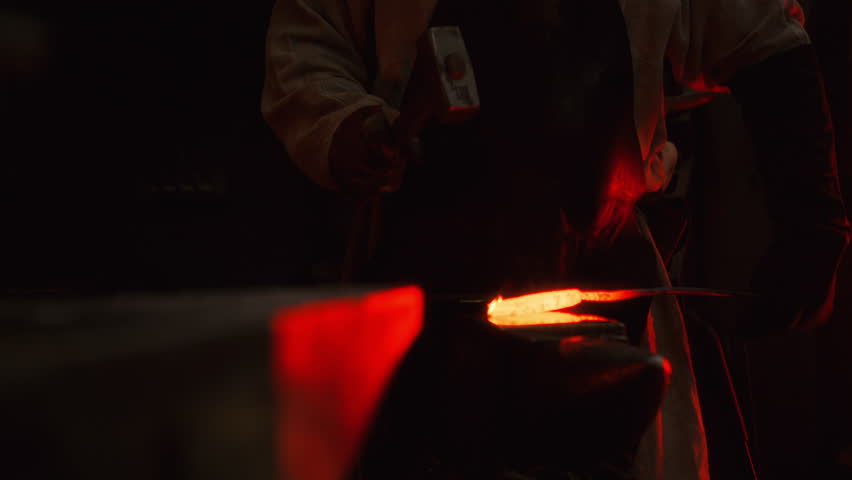 A blacksmith uses a hammer to shape a piece of red-hot metal in a forge on an anvil, creating a future product, sparks fly from blows. The process takes place in the darkness of the forge.