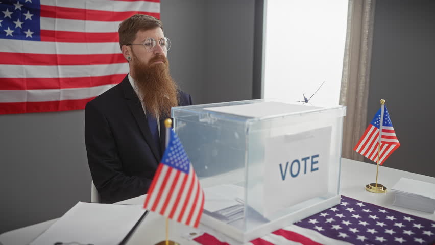 A bearded man in a suit smiles near a us voting ballot box with american flags indoors.